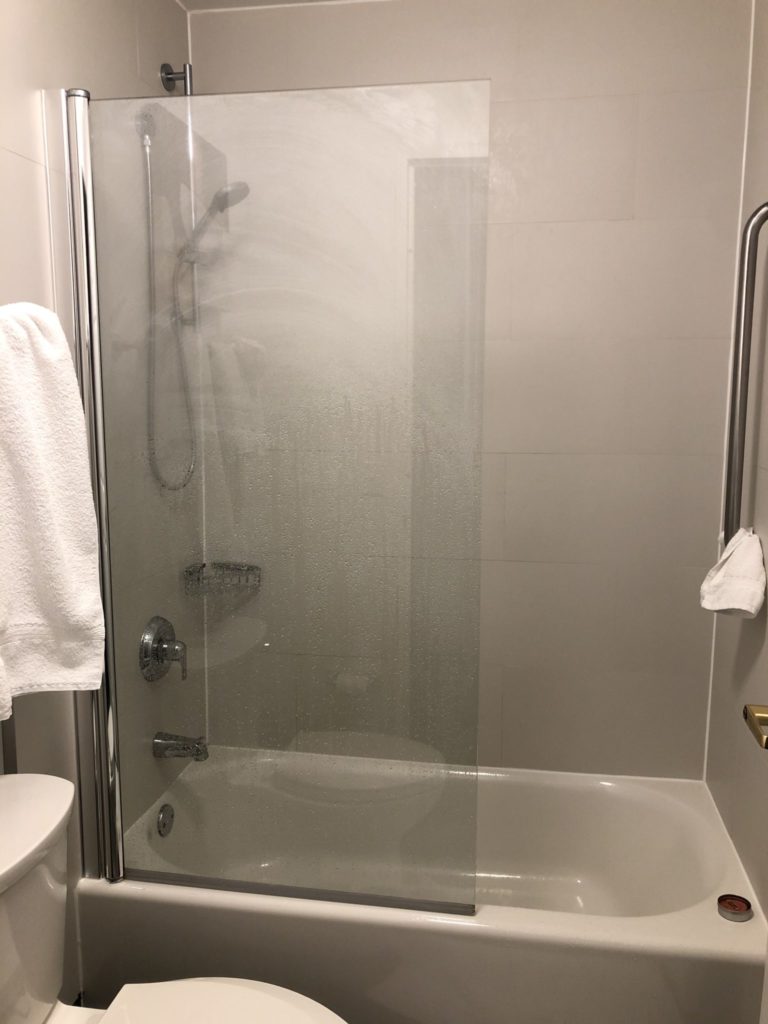 A hotel bathroom. From the door, the toilet is on the left. The tub is on the back wall behind the toilet. A glass panel covers half the shower area to ensure the toilet is not soaked. The shower controls can be seen (barely) through the wet glass. 