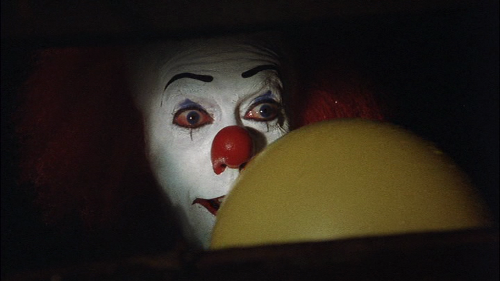 Tim Curry as the clown in Stephen King's It, holding a yellow ballon in a storm drain, looking overly friendly