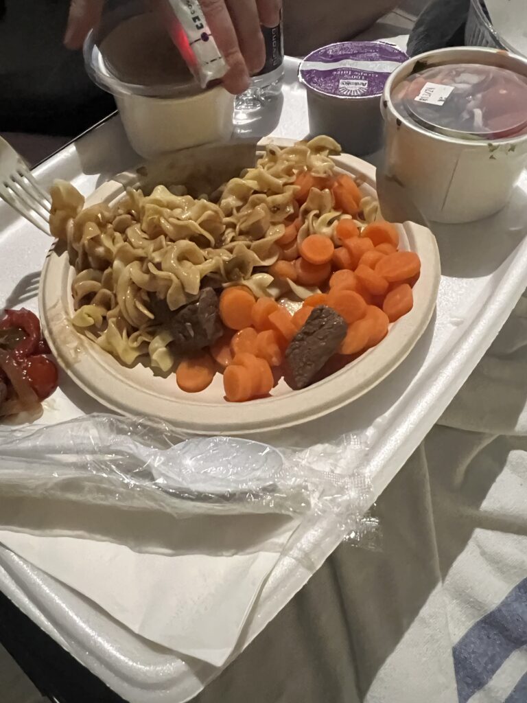 A hospital tray with a plate loaded with egg noodles, vegetables, and beef. other containers hold salad and apple crisp.